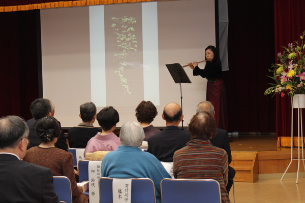表彰者が椅子で座って舞台で楽器を演奏されている女性を観覧している写真