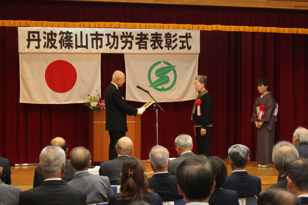 功労者表彰式典舞台上で市長から表彰を受けている二名の女性が並んでいる様子の写真