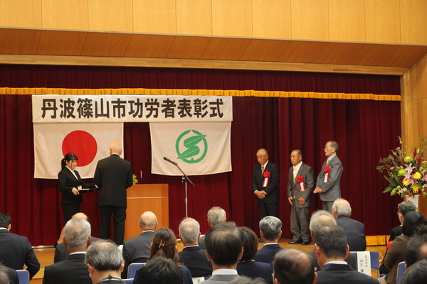 功労者表彰式典舞台上で市長から表彰を受けている三名の男性が並んでいる様子の写真