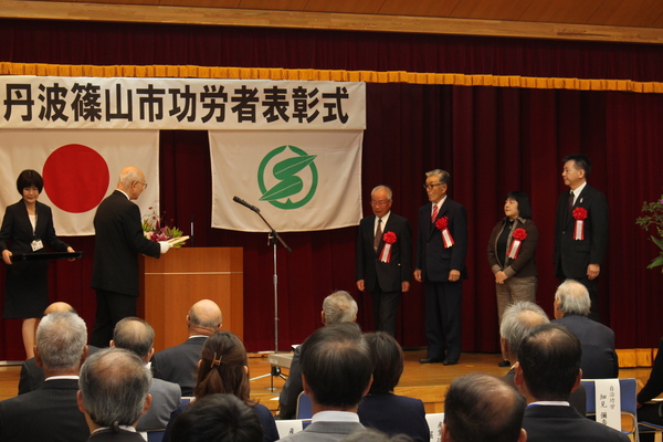 功労者表彰式典舞台上で市長から表彰状を読み上げ、四名の男性女性が聞いている様子の写真