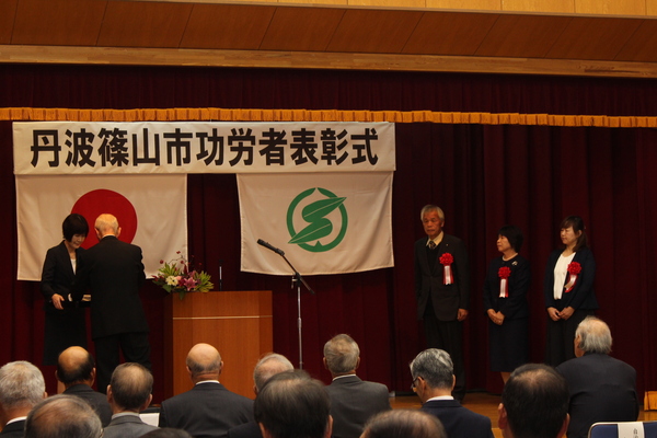 功労者表彰式典舞台上で市長から表彰を受けている三名の男性女性が並んでいる様子の写真