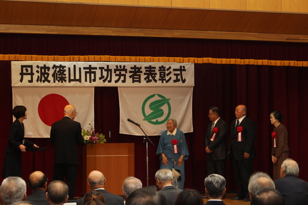 功労者表彰式典舞台上で市長から表彰を受けている四名の男性女性が並んでいる写真
