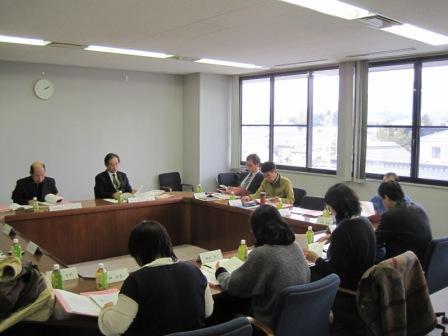机の上にお茶と資料を用意し会議室内で会議を行う委員たちの写真