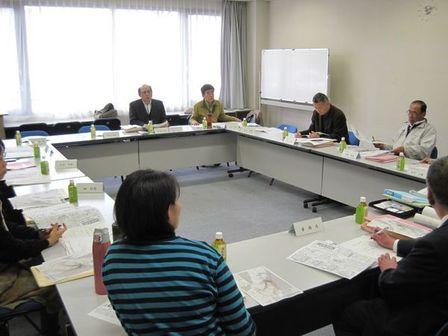 ホワイトボードと広い窓のある会議室で会議を行う委員たちの写真
