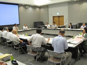 スクリーンのある会議室内でマイクを使い発言する委員と耳を傾ける委員たちの写真