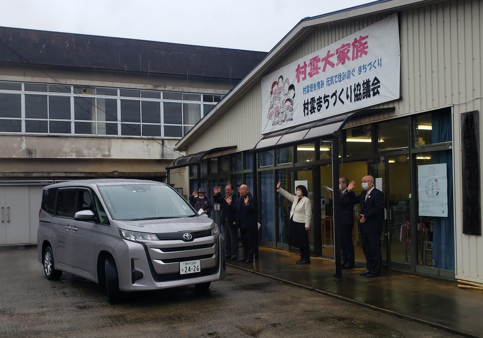 有償運送の車両が出発し、関係者が手を振って見送っている