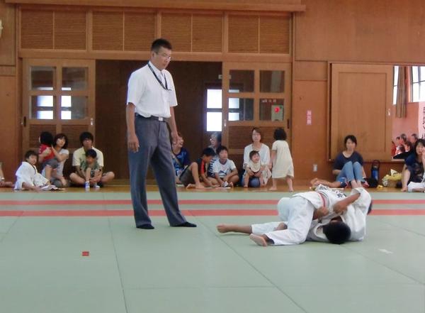 畳の上で柔道をしている2人の少年とその横に審判、その後ろに応援する人たちが映っている写真