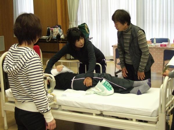 ベッドに寝ている人の脇と足に女性が手をかけながら、ベッドの足元に立っている女性に説明をしている写真