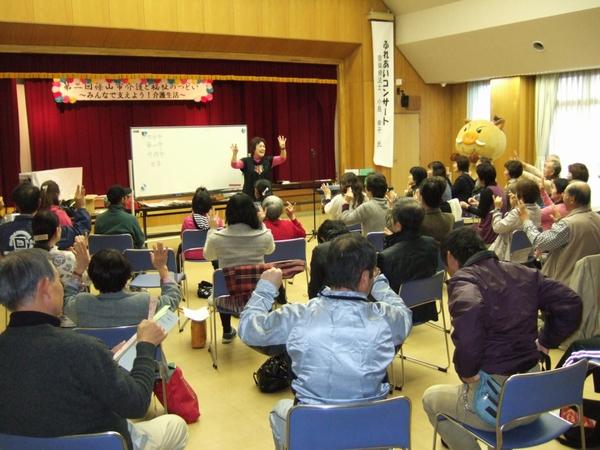 ふれあいコンサートと幕がかかっている横で女性が手をあげて話をしていて、前に座っている人達も一緒に手をあげている写真