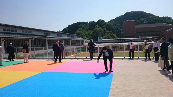カラフルなマットがある屋外で10名ほどの人達が立って見学している写真
