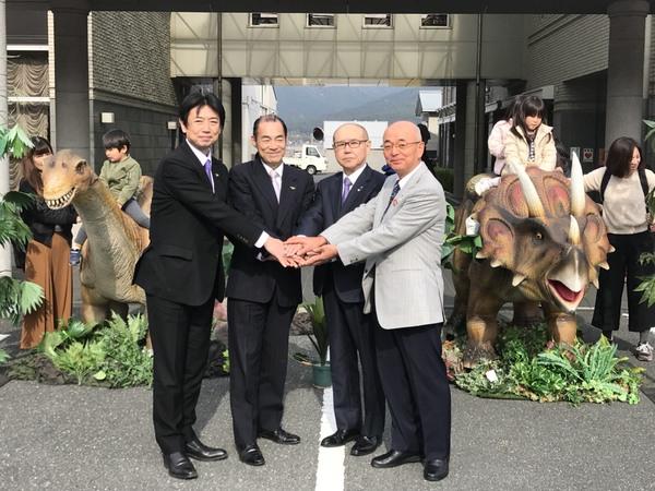 2体の恐竜の模型にまたがっている子どもたちの前で市長と関係者3名が手を前に出し4人で手を重ねている写真