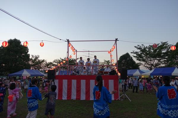 提灯で飾られた舞台の上で演奏をしている方々を見ながら輪になり踊りを踊っている人々の写真