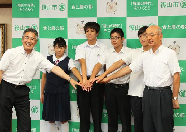 降矢信さん、 野々口慎也さん、 溝畑樹蘭さん、 谷本藍子さんの4人と、市長、男性で手を伸ばし重ねている写真