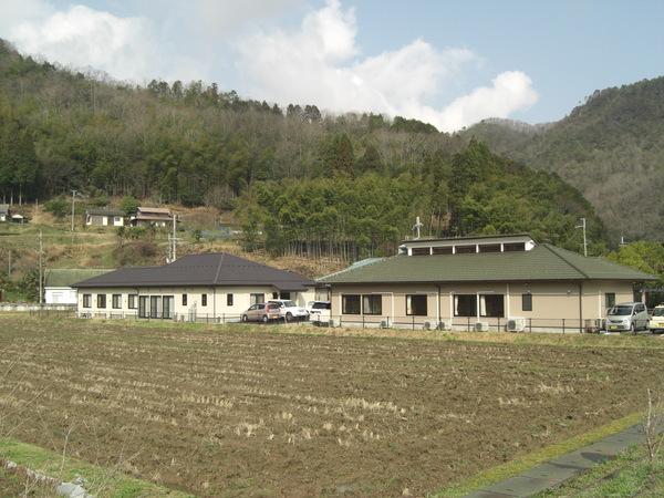 山を背景にしたのどかな風景に開設された緑色の屋根とこげ茶色の屋根の2棟の幸の郷の写真