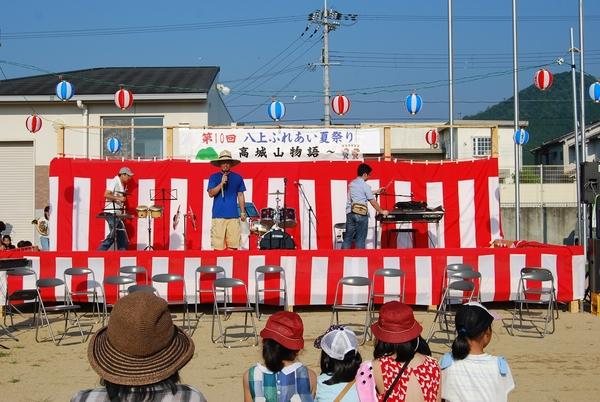 八上ふれあい夏祭りのステージで麦わら帽子を被った男性が演奏に合わせて歌を歌っている様子を見ている人々の写真