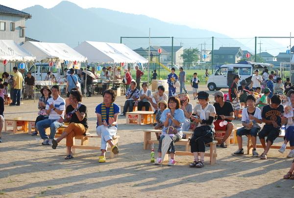 ステージの出し物を校庭に設置された椅子に座って見ている人々の写真