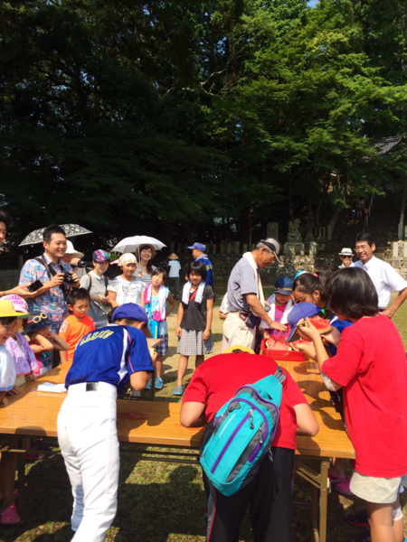 長テーブルでスイカを食べている子供たちがおり、その様子を見ている人々の写真