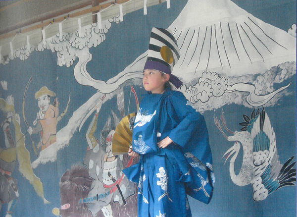 黒と白のシマシマに金色の丸印がついて、紫色のハチマキをで締めた烏帽子を被り、青色の狩衣の衣装を着た男の子が金色の扇子を持って腰に手を当てて立っている「追入神社の子ども三番そう」の上演の様子の写真