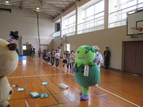 児童たちが両手を胸の前で交差して、ゆるきゃら「まるいの」「まめりん」の後に続いて、歩きながら踊っている様子の写真