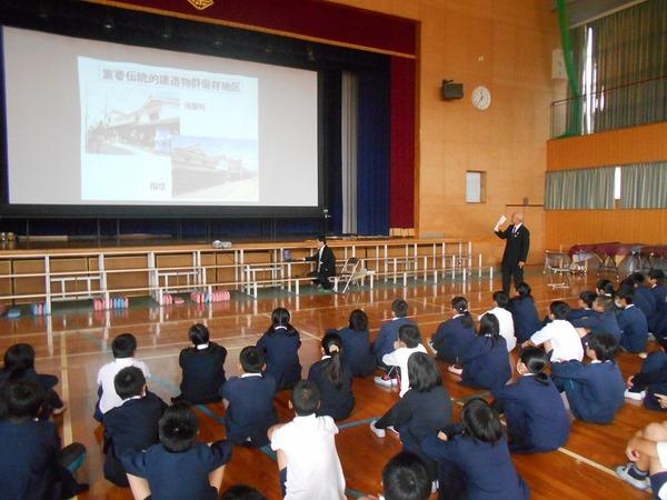 体育館のスクリーンに映っている映像を見ながら、市長が床に座っている児童達に話をしている様子の写真