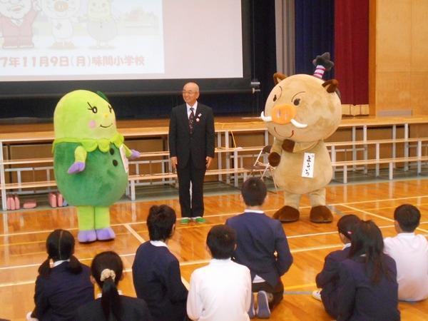 体育館で児童達が座っている前に、ゆるきゃら「まめりん」、市長、「まるいの」が一列に並んで立ち、児童に話をしている写真