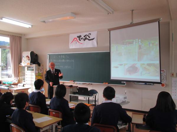 教室の教壇に立った市長が映し出されているスクリーンの映像の横で生徒たちに説明している写真