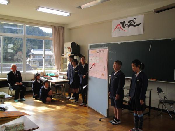 生徒が作成した作品に指差し棒を向けて説明しているのを市長が窓際の椅子に座り聞いている写真