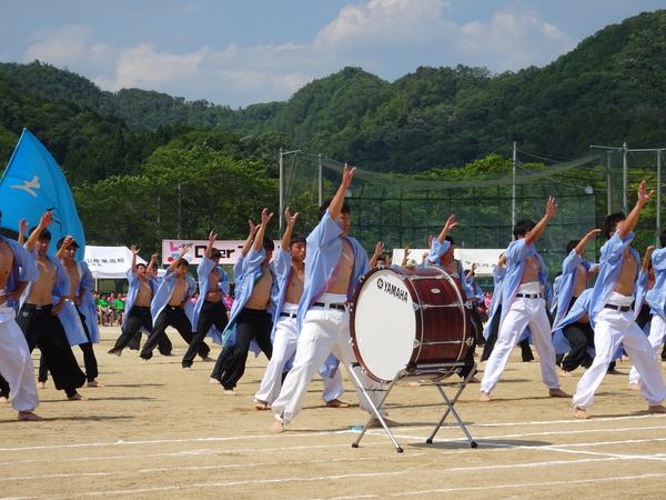 薄い紫の法被をきて、黒色と白色の長いズボンを履いた男子生徒たちが、片手をあげている写真