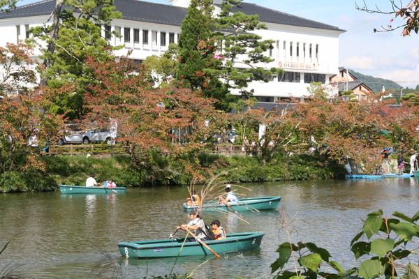 白い建物と色々な木々の前のお濠で緑色の3隻のボートで観光している人達の写真