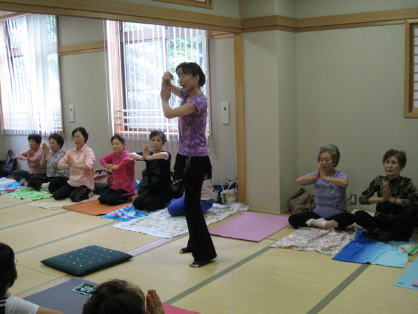 畳の部屋の中央にヨガの指導をする女性が立っていて、その周りにはタオルをひいた上に座って手で拝むようなヨガのポーズをしている女性たちの写真