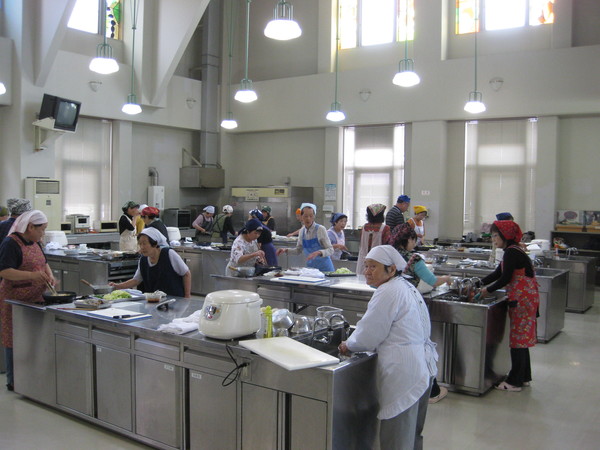 7台ほどの調理台の上に炊飯器、まな板、包丁などが置かれていて、エプロン姿の女性たちが作業をしている写真