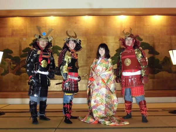 金屏風の前で着物姿の女性が1人、鎧兜姿の男性が3人並んでいる写真