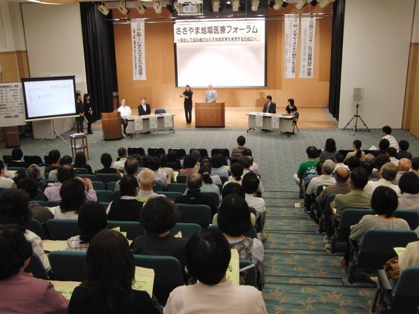 ささやま地域医療フォーラムと書かれた幕とスクリーンの前に教卓があり、そこに男性が立っていて、たくさんの座っている参加者達に話をしている写真
