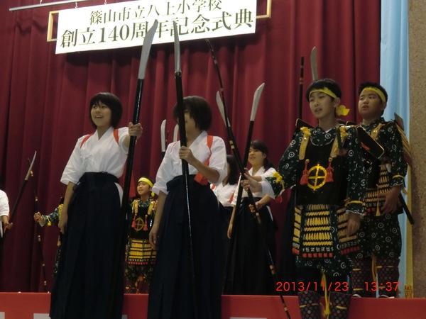 袴を着て、槍を持っている女子児童と、甲冑を身にまとい、槍を持っている男子児童が舞台の上に立っている写真