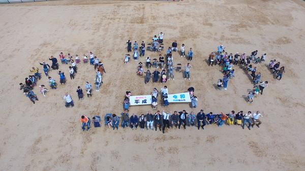 「のなか」と人文字でつくっていて平成と令和がかかれたものを手にもつ人達がはっきりわかる高さで写した空からの写真