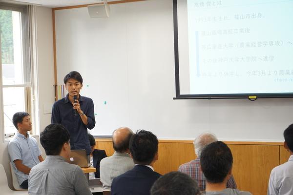 高橋 俊さんがマイクを持ってスクリーンに映し出されている内容について話しているのを聞いている人々の写真