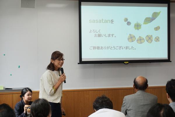 渋谷 啓子さんがマイクを持ってスクリーンに映し出されている内容について話しているのを聞いている人々の写真