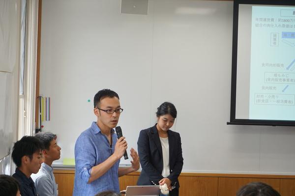 西川 勢二郎さんがマイクを持ってスクリーンに映し出されている内容について話しているのを後ろで立って聞いている女性と横で椅子に座って聞いている男性らの写真