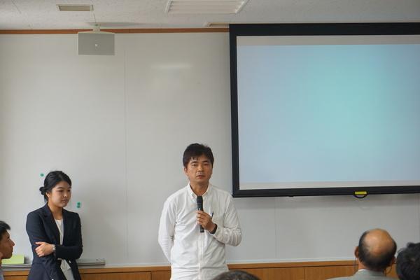 橋本 豊彦さんがマイクを持って話しているのを後ろで立って聞いている女性と座って聞いている人々の写真
