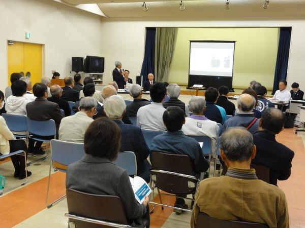 プロジェクトスクリーンにに注目する会議参加者と、マイクをもって話す男性の写真