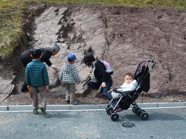 スーツを着た男性が地面の断面を指でさし、子供連れの家族がそれを見ている写真