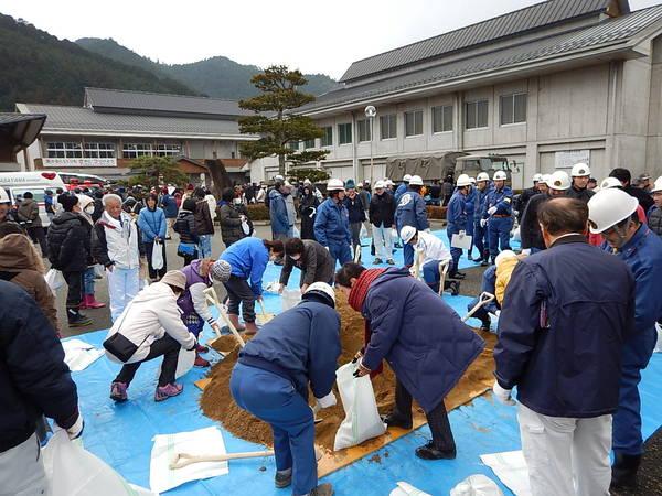 ブルーシートの上に沢山の土を置き、地域の方がスコップを使って白い袋に入れ土嚢を作成している写真