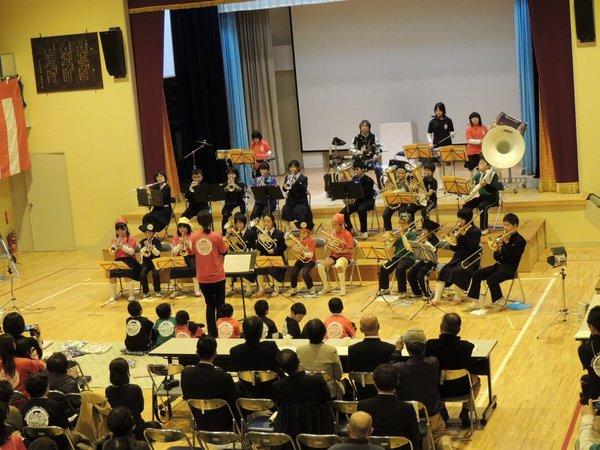 体育館の舞台の前で、吹奏楽の生徒達が演奏しており、後方に沢山のお客さんが椅子に座って演奏を聴いている様子の写真