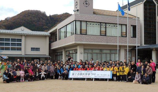 大芋小学校の校舎の前で、「第10回大芋まつり」と書かれている看板の後ろに、参加者達が集まっている集合写真