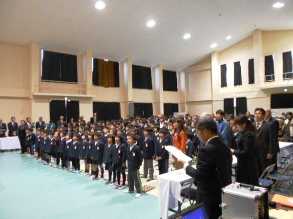 体育館で観客席の児童達も整列して歌を歌っており、両脇にいる人も立ち上がって一緒に歌を歌ったり、手元の紙を目で追っている様子の写真