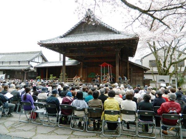 屋根のある舞台で行われている能をたくさんの人達が椅子に座って観劇している写真