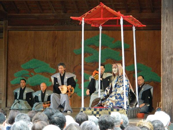 舞台の中央に能のお面をかぶった人が座っていて、その横にお囃子の男性二人が小太鼓を叩いている写真