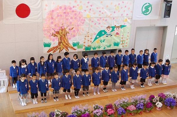 後ろの壁に桜の木やつくし、子供の絵が描かれており、ステージ上に青の上着を着た新入生が立っている所を上から撮影した写真