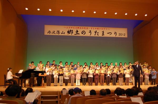 「丹波篠山 郷土のうたまつり 2012」の垂れ幕のある舞台上で、ピアノの伴奏に合わせて、合唱団の方が歌詞を見ながら歌を歌っている様子の写真