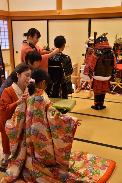 畳の部屋で、小さな女の子や少年たちが着物や鎧兜を大人達から着せてもらっている写真